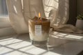 Candle in a glass jar with a label on a light surface, draped with sheer curtains.